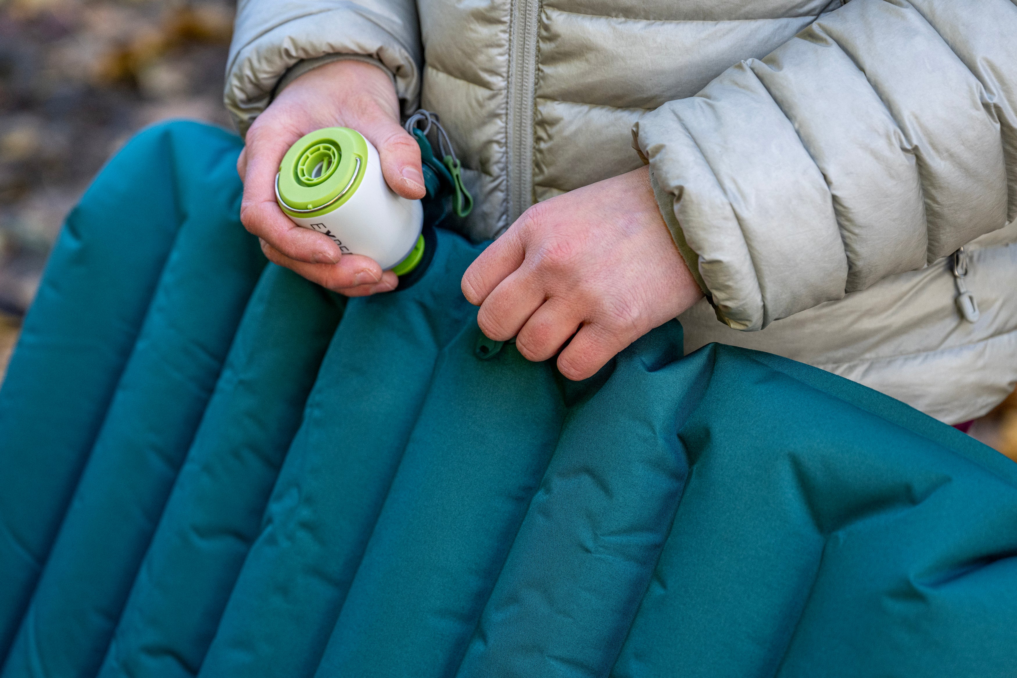 A person in a gray puffer jacket is inflating a teal camping mattress with a small, white and green portable pump. The scene conveys outdoor preparedness.